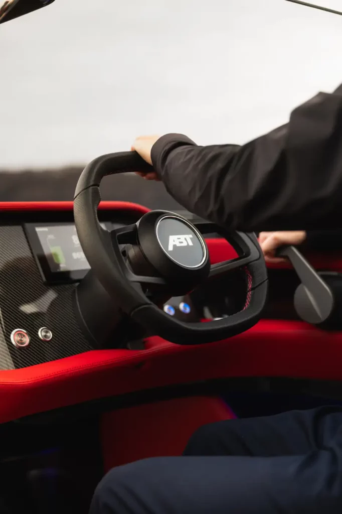 Detailansicht des Cockpits der ABT Marian M 800-R, fokussiert auf eine Hand, die den modernen Schalthebel bedient, eingebettet in eine Umgebung aus Karbonfaser und rotem Leder.