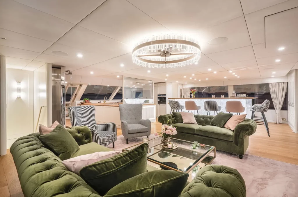 A spacious and luxurious living area on the 100 Sunreef Power yacht, with green velvet sofas, striped armchairs, a large chandelier, and a view of the open kitchen in the background.
