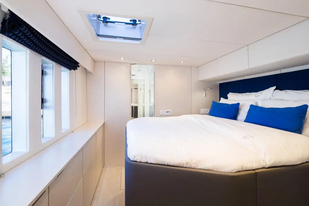 A cozy bedroom on the Sunreef 50 yacht with a large bed dressed in white linens and blue pillows, with natural light streaming in through side windows.