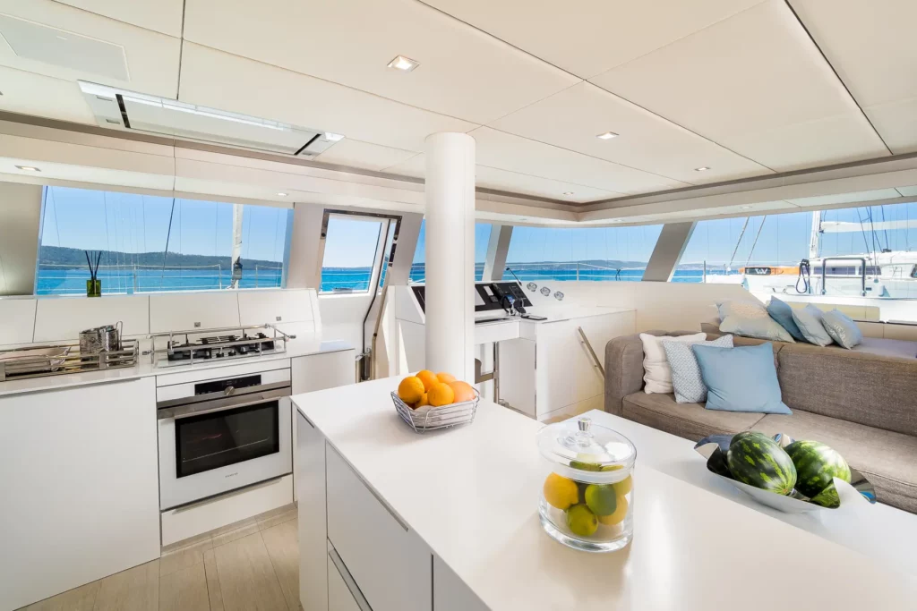 A sleek, modern kitchen on the Sunreef 50 yacht, featuring white countertops, a central island with fruit baskets, stainless steel appliances, and large windows providing a view of the sea.