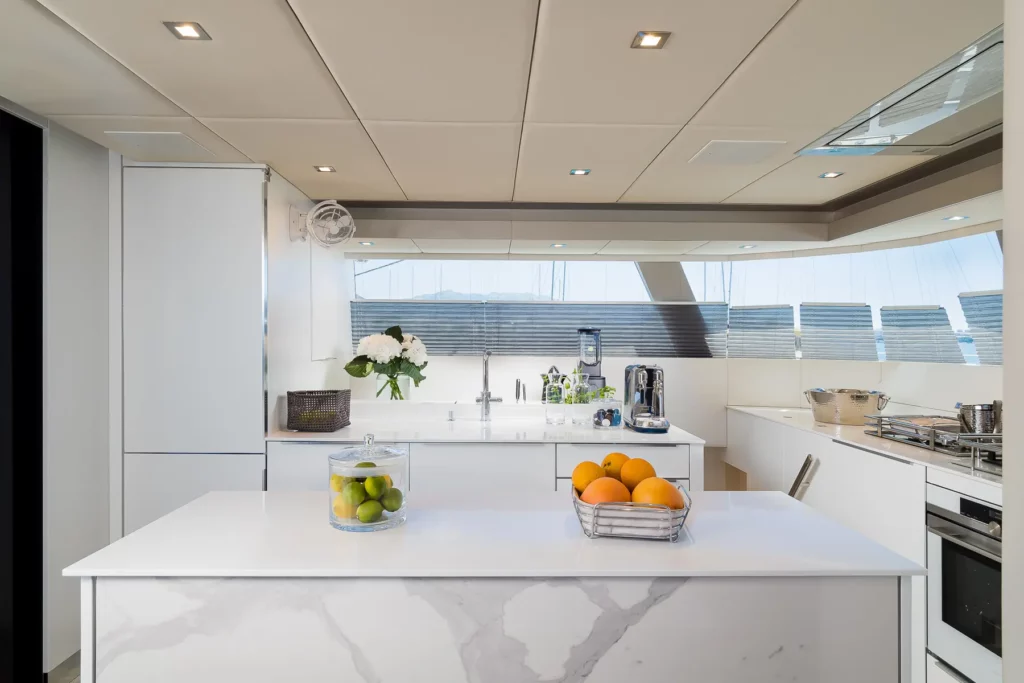 A sleek, modern kitchen on the Sunreef 50 yacht, featuring white countertops, a central island with fruit baskets, stainless steel appliances, and large windows providing a view of the sea. The space is bright and airy, with elegant decor including a vase of fresh flowers and minimalist design elements.