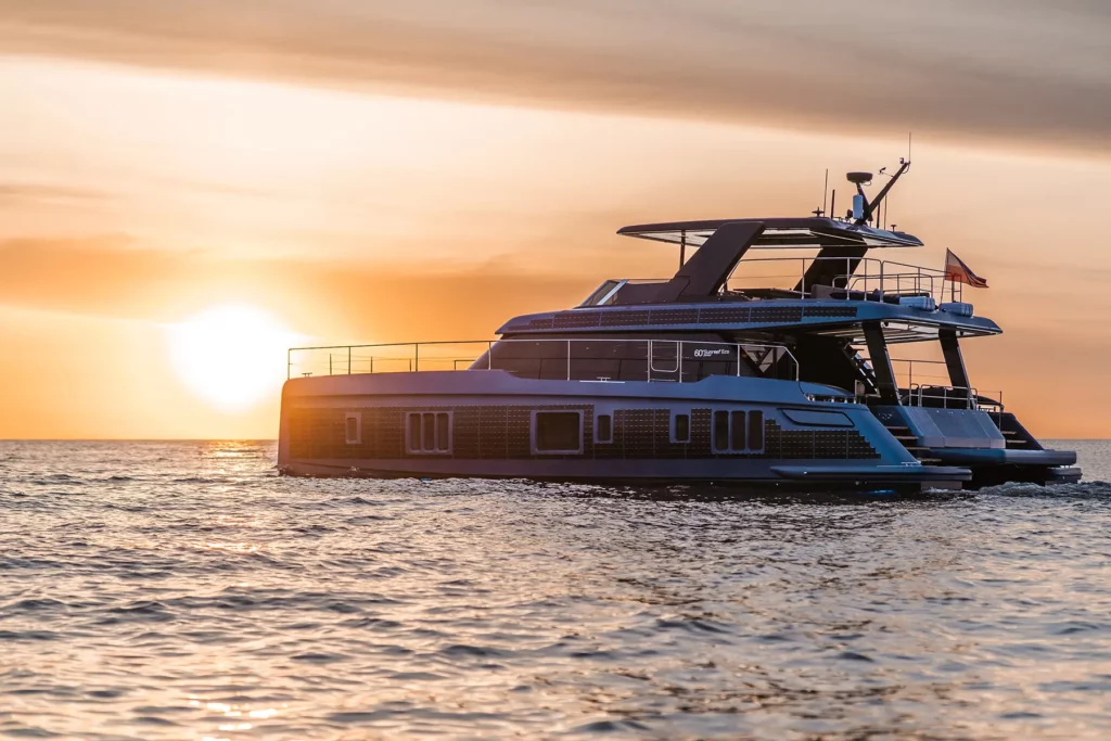 Die Sunreef Power 60P Eco Yacht fährt bei Sonnenuntergang auf offenem Meer. Die Silhouette der Yacht vor dem orangefarbenen Himmel schafft ein Bild von Luxus und Nachhaltigkeit.
