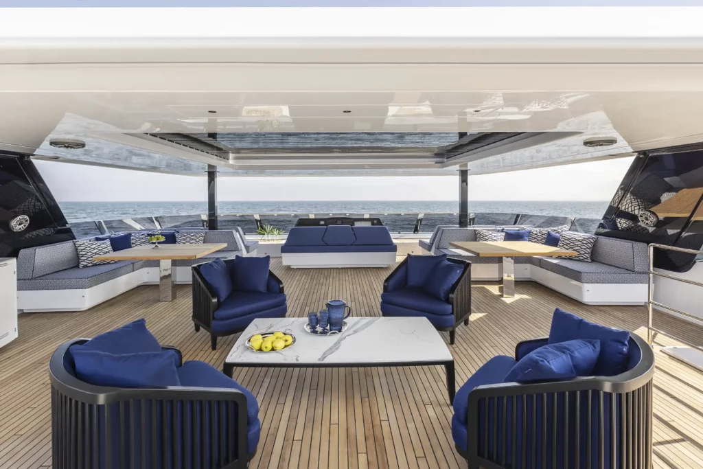 Die geräumige Flybridge der 80 Sunreef Power, ausgestattet mit eleganten blauen Polstern und modernen Sitzgelegenheiten, bietet eine perfekte Lounge-Atmosphäre für das Genießen der Aussicht auf das Meer.