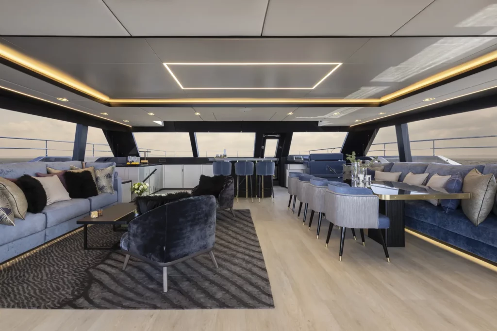 Ein luxuriöser Lounge-Bereich auf der 80 Sunreef Power Yacht, mit plüschigen blauen Sofas, die mit gemusterten und Samtkissen verziert sind, einem modernen Essbereich mit gepolsterten Stühlen und einer eleganten, minimalistischen Bar mit hohen Hockern, alles umgeben von großen Fenstern, die einen Panoramablick auf den Ozean bieten.