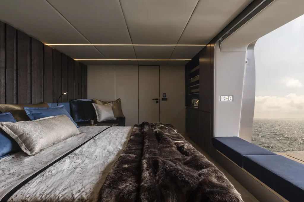Ein luxuriöses Hauptschlafzimmer auf der 80 Sunreef Power Yacht, mit einem großen Bett, plüschiger Bettwäsche und Kissen, warmer Beleuchtung und einer Fensterwand, die sich zu einem privaten Deck mit atemberaubendem Meerblick öffnet.