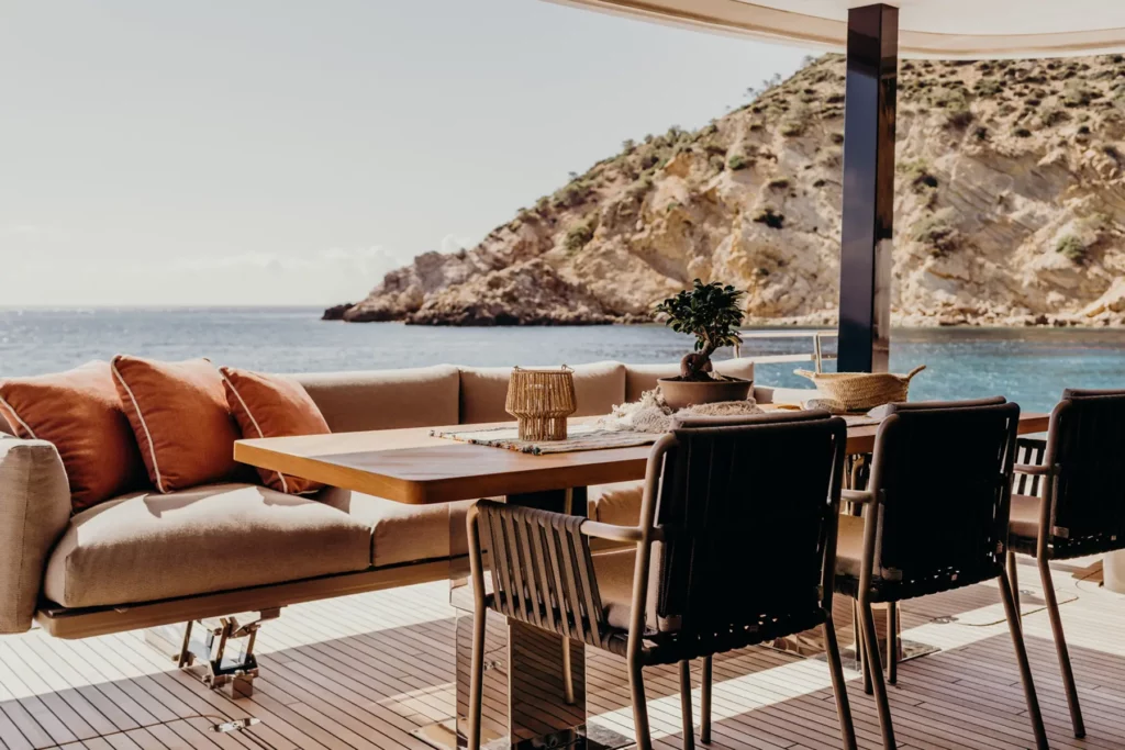 Eleganter Essbereich im Freien auf dem Achterdeck des Sunreef 80 Katamarans, mit bequemen Sitzgelegenheiten und Blick auf eine felsige Küste.