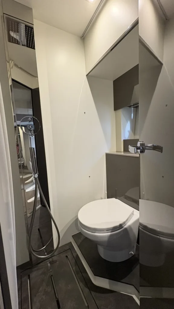 Kompaktes Bad der Cranchi M44 mit WC, Dusche/Handbrause, Duschvorhang und Ablagen in hellem Interieur.