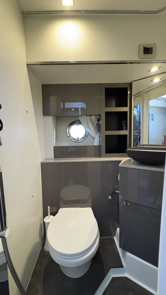 Badezimmer der Cranchi M44 frontal mit WC, grauen Hochglanz-Schrankelementen, Bullauge und Waschtisch rechts.