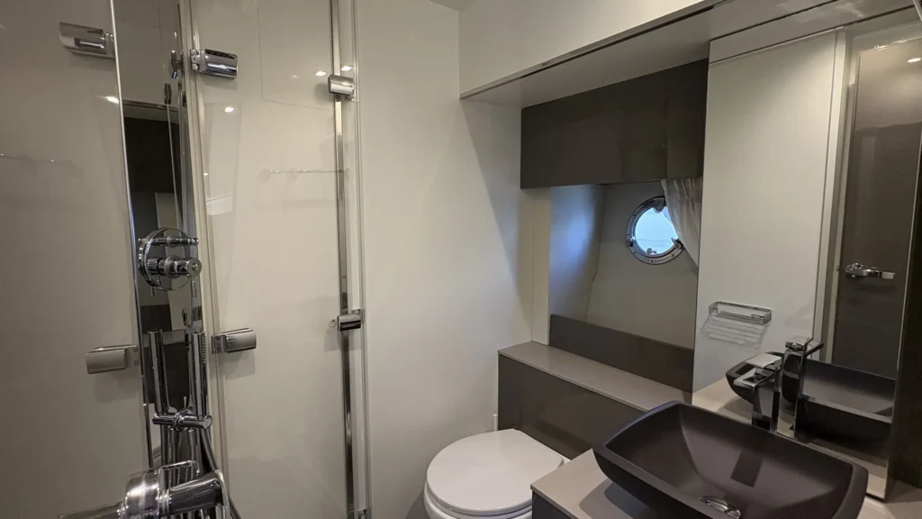 Bad der Cranchi M44 mit Duschkabine links, WC, Waschtisch mit dunkler Schale und Spiegel, Bullauge im Hintergrund.